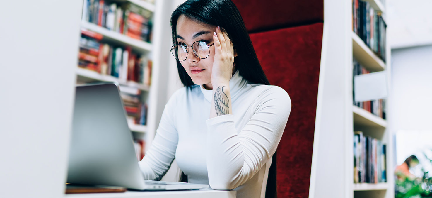 Asiatische Frau mit Brille sitzt am Laptop in einer Bibliothek.