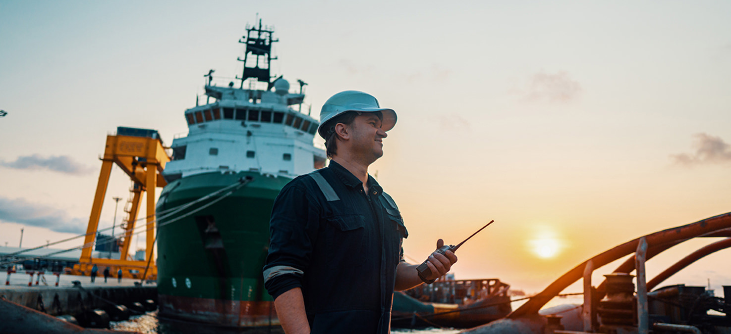 Mann mit Helm und Funkgerät steht vor einem großen Frachtschiff im Hafen.